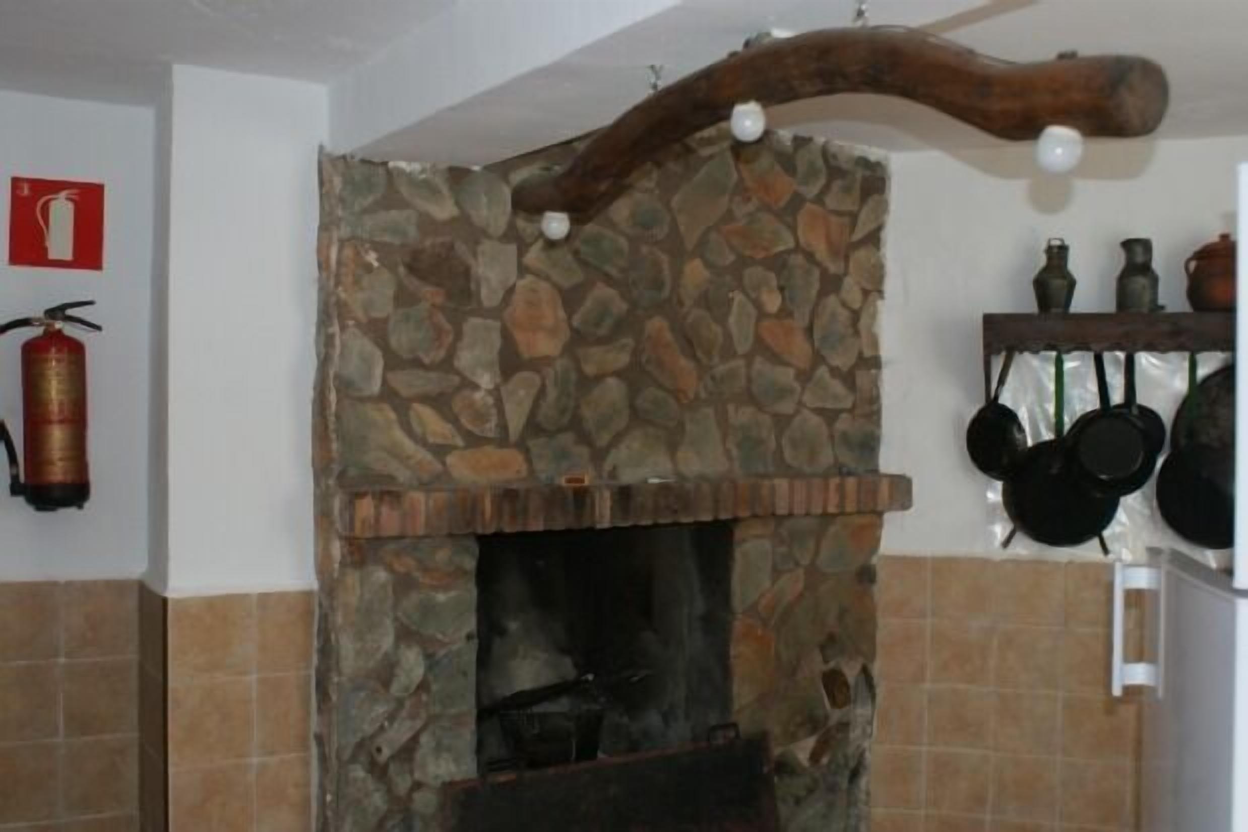 Un camino in pietra con una trave in legno rustica sopra, un estintore a parete e pentole appese.