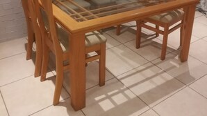 Ein rechteckiger Esstisch mit Glasplatte, umgeben von sechs hölzernen Stühlen, jeweils mit gepolsterten Sitzflächen. Der Tisch steht vor einer weißen Ziegelwand, der Boden ist mit Fliesen belegt.