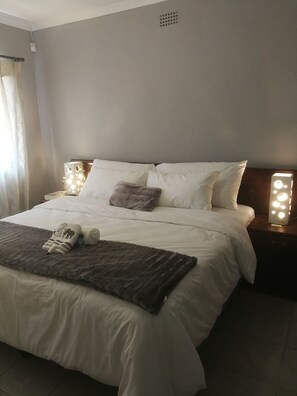 A neatly made bed with white linens, a brown throw blanket, and a pair of shoes on the bed. There are two bedside tables with illuminated lamps. - Cape Pine Guest House (Cape Town)