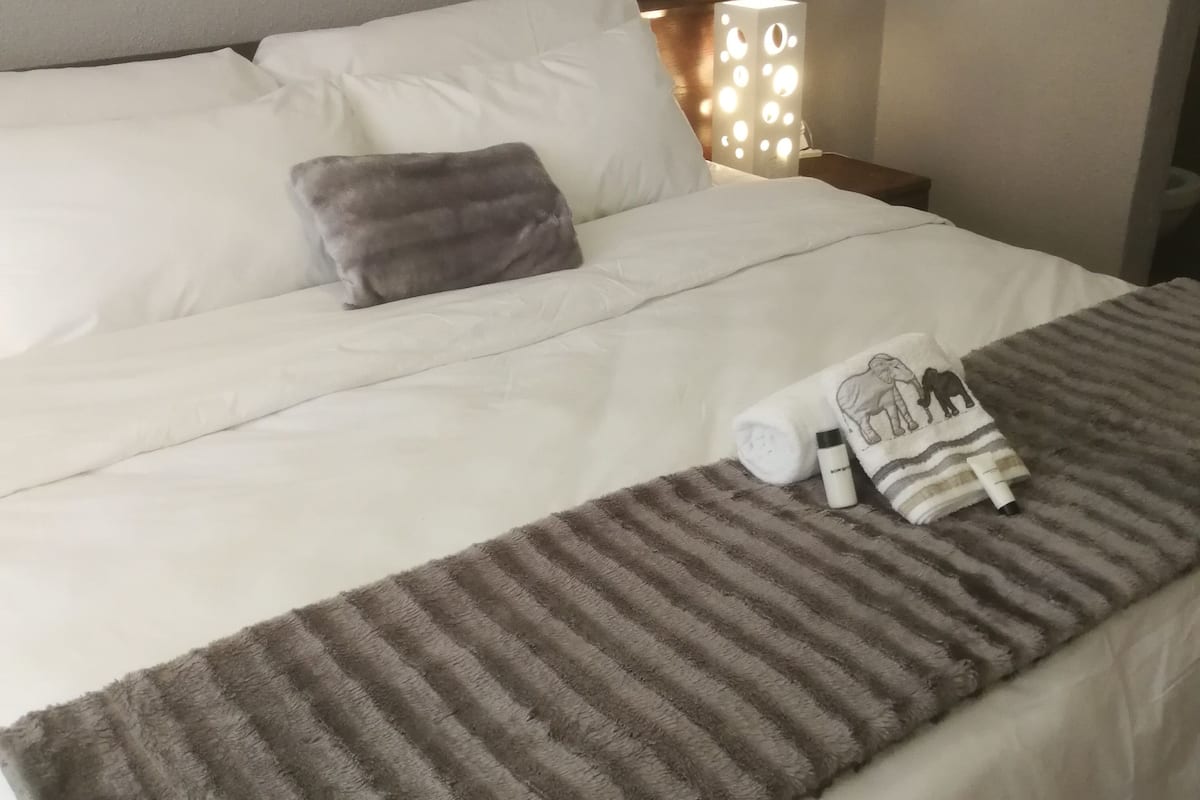 Una cama bien hecha con sábanas blancas, una manta gris a rayas y una toalla enrollada con estampado de animalitos.