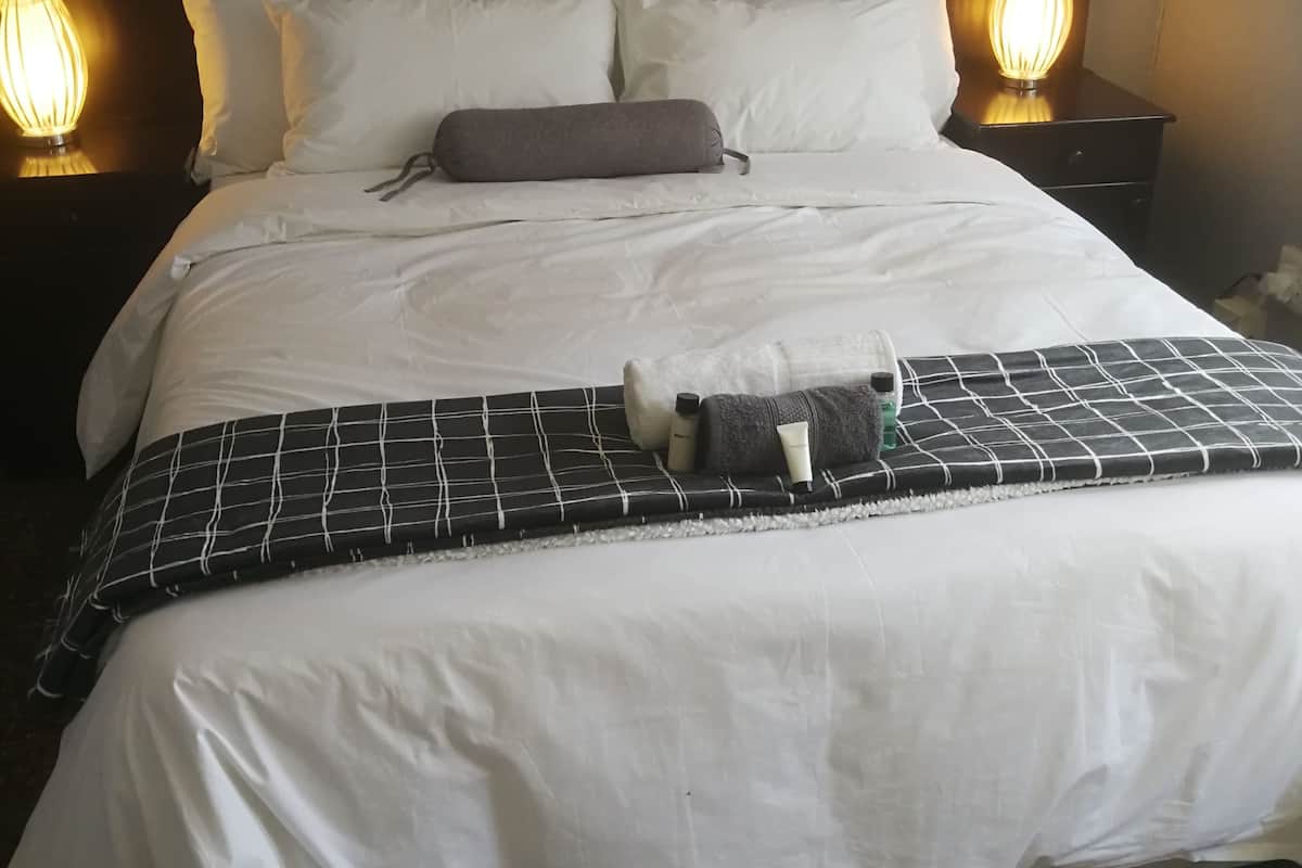 Una cama bien hecha con sábanas blancas, una manta de cuadros blancos y negros, y una almohada. Hay dos lámparas de noche a cada lado de la cama.