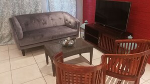 Ein Wohnzimmer mit grauem Sofa, einem Glastisch, zwei Holzstühlen, einem Fernseher auf einem Holzboden und einer roten Backsteinwand.