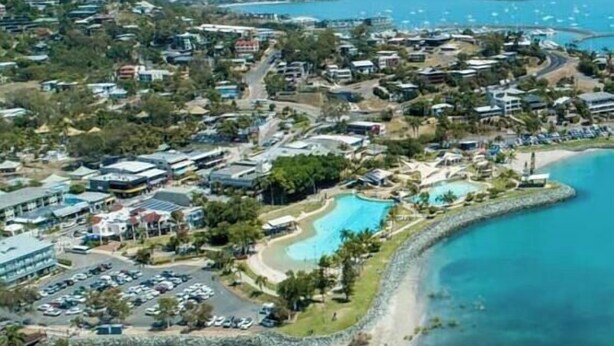 Vista aerea di una località balneare con una grande piscina, spiaggia e una zona residenziale densa con strade e edifici.