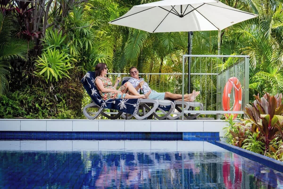 A couple relaxing by a poolside lounge chair with a white umbrella, surrounded by lush tropical plants.