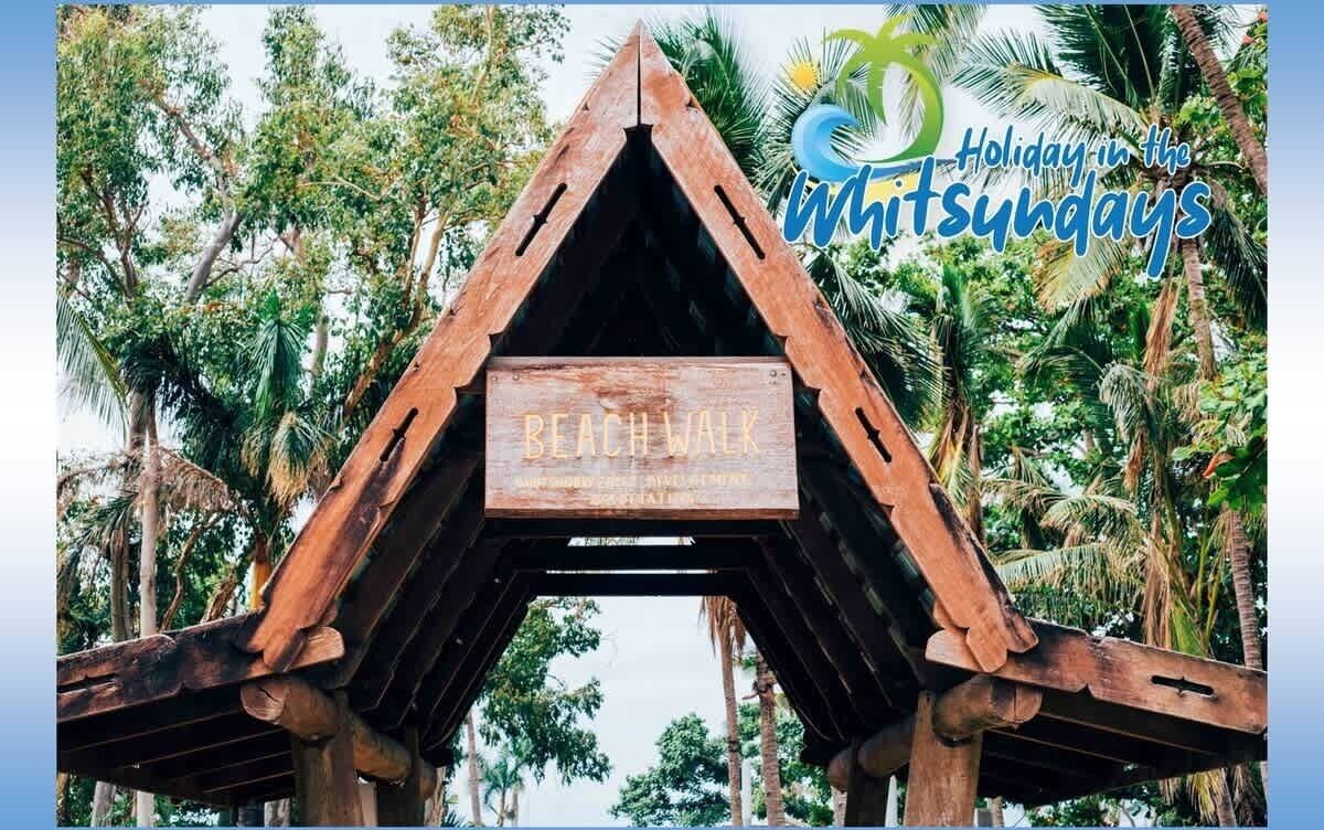 Un panneau en bois portant l’inscription « PROMENADE SUR LA PLAGE », placé dans une structure triangulaire et entouré de palmiers.