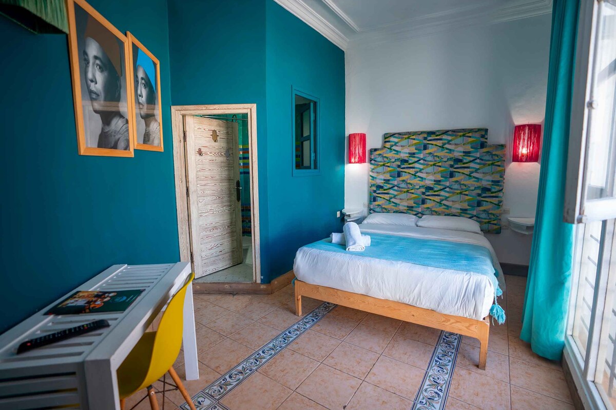 A bedroom with a bed, a desk, and a chair. There is a door leading to another room.