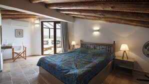 A bedroom with a large bed, wooden ceiling beams, a window with curtains, and a small table with a lamp.