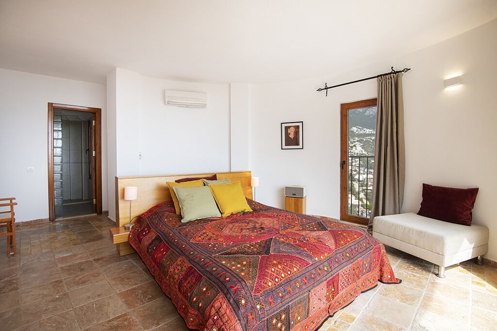 A bedroom with a large bed, a bedside table, a nightstand, a window with curtains, and a view of the outdoors.