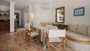 A dining area with a round table, four wooden chairs, and a bowl of fruit. There is a sofa with cushions and a window with a view of trees.