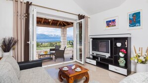 A living room with a sofa, coffee table, TV, and a balcony with a view of mountains. - Villa Agape - Three Bedroom Villa with Swimming Pool (Mirca)