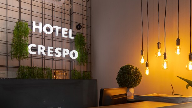 En receptionsdisk med et skilt der siger "HOTEL CRESPO", en potteplante og hængende lyspærer.