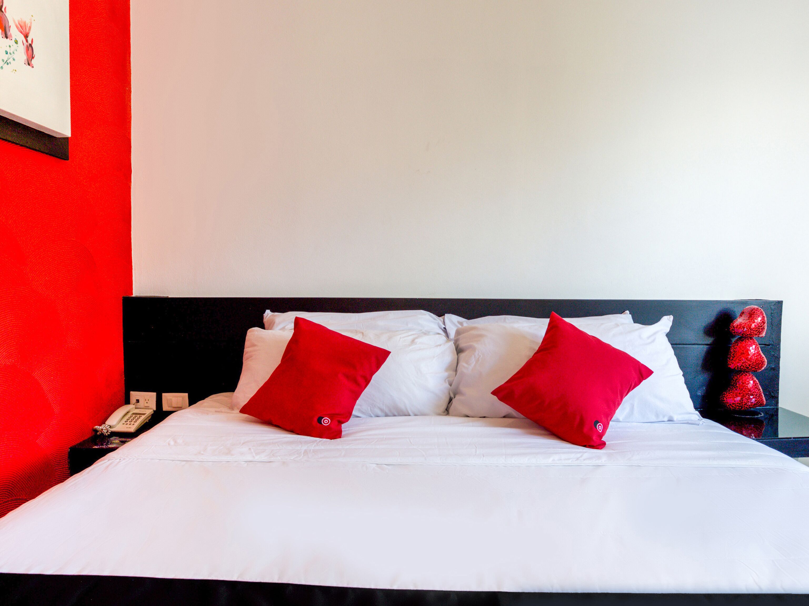 A hotel room with a bed, white bedding, two red pillows, a phone on a bedside table, and a red wall.