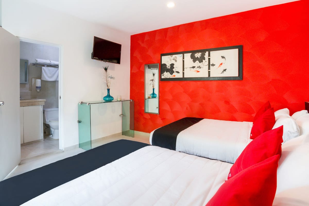A bedroom with a red accent wall, a bed with white and red bedding, a small bedside table with a vase, and a mounted TV.