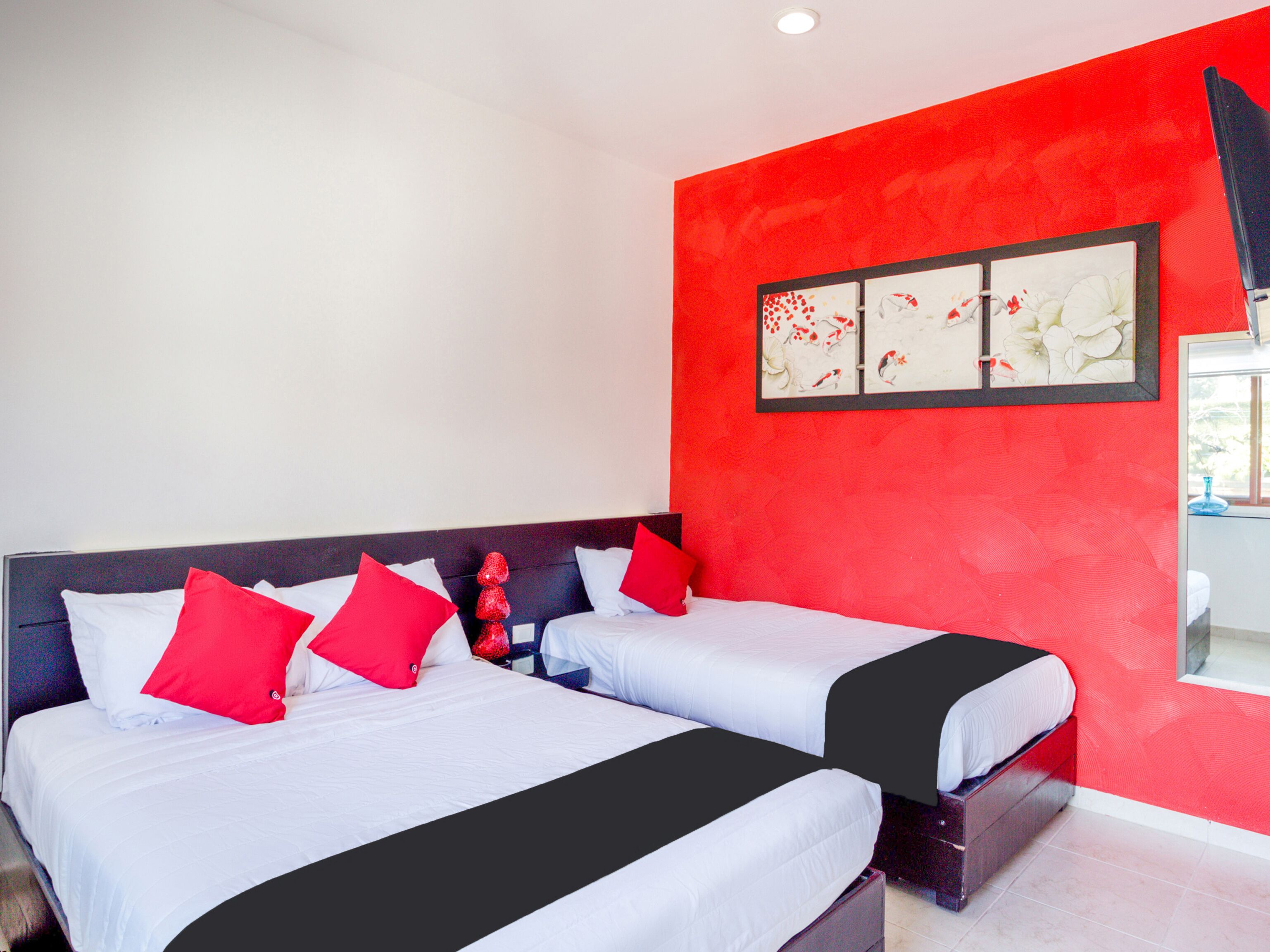 A hotel room with two beds, red and white bedding, a red accent wall, and a framed artwork above the beds.
