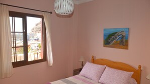 A bedroom with a bed, a painting, a window with curtains, and a view of a building outside. - Accommodation Junonia Marítimo 1 (Playa de Santiago)