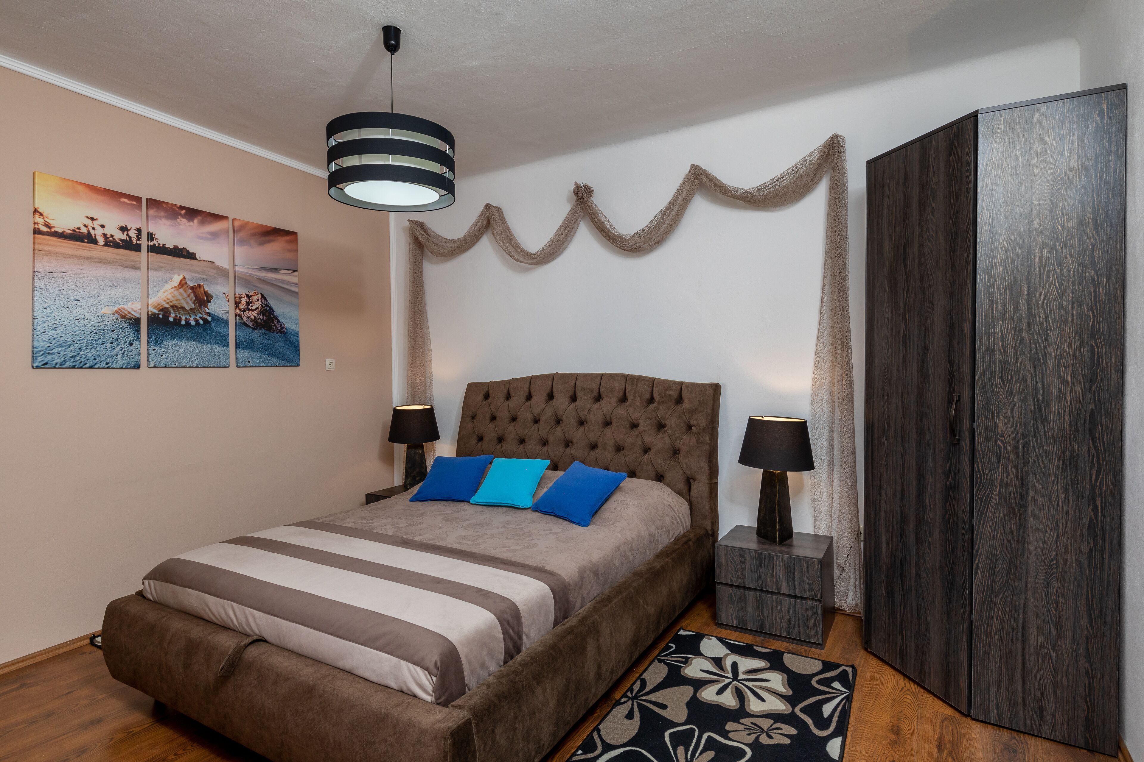 A bedroom with a large bed, two bedside tables, a wardrobe, a hanging light fixture, and a wall-mounted artwork.