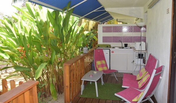 A small kitchen with a sink, a microwave, and a refrigerator. There is a wooden table with a chair and a small table with a pink bowl. The kitchen is adjacent to a wooden deck with a blue awning.