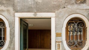A building entrance with a green door, a tiled floor, and two ornate windows with metal bars.