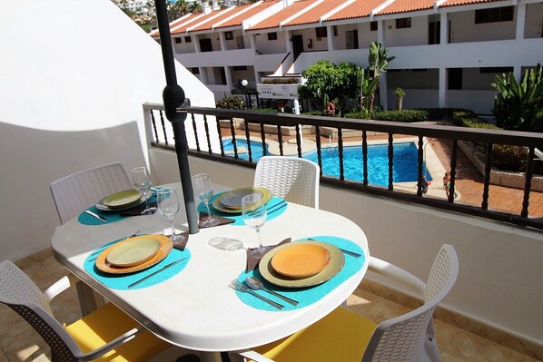 Une table à manger dressée avec des assiettes, des couverts et des verres, donnant sur une piscine et un bâtiment dont le toit est recouvert de tuiles.