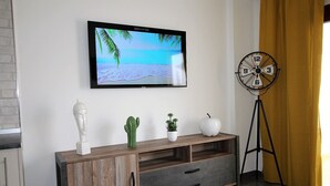 A modern living room with a flat-screen TV on the wall, a wooden console table with decorative items, a floor lamp, and a window with yellow curtains. - Apartment with large terrace and beautiful pool view, solarium, private parking (Costa Adeje)