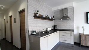 A modern kitchen with white brick walls, a black countertop, and white cabinetry. There is a wooden door on the left, a shelf with plants, and a coffee maker on the counter. - Apartment with large terrace and beautiful pool view, solarium, private parking (Costa Adeje)
