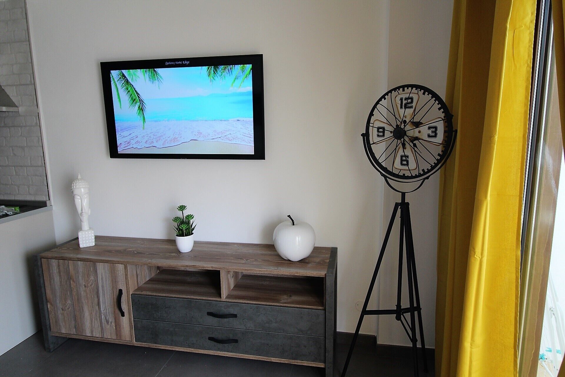 Una habitación con un armario de madera, una pequeña planta en maceta, un jarrón blanco, un televisor empotrado en la pared mostrando una escena de playa y un reloj grande sobre un pedestal.