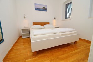 Une chambre avec un plancher en bois, un lit blanc avec deux oreillers, une table de chevet, une lampe fixée au mur et une fenêtre avec des stores.