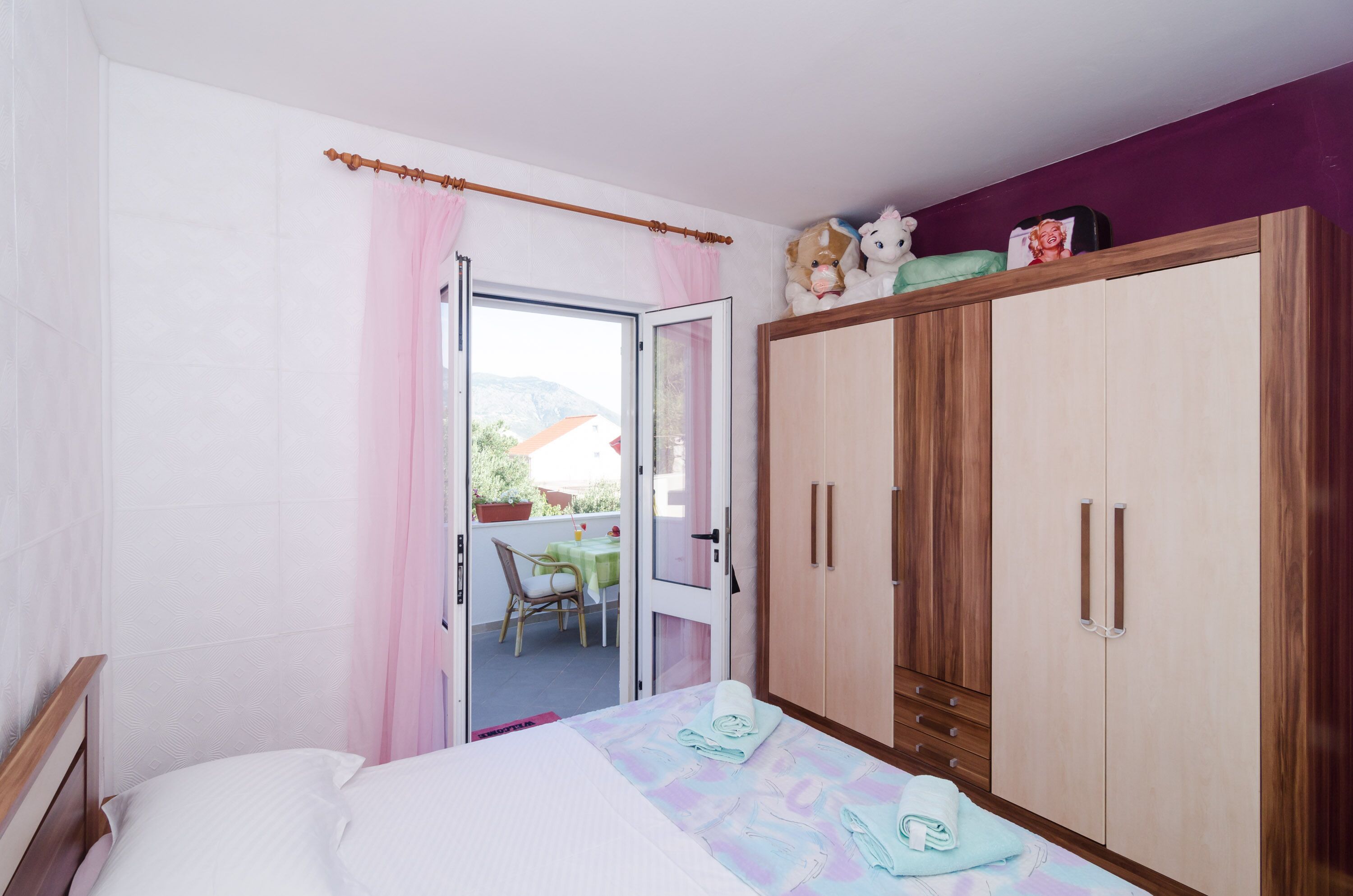 Une chambre avec un lit, une armoire en bois, un balcon avec une table et une chaise, et une vue sur l’extérieur.