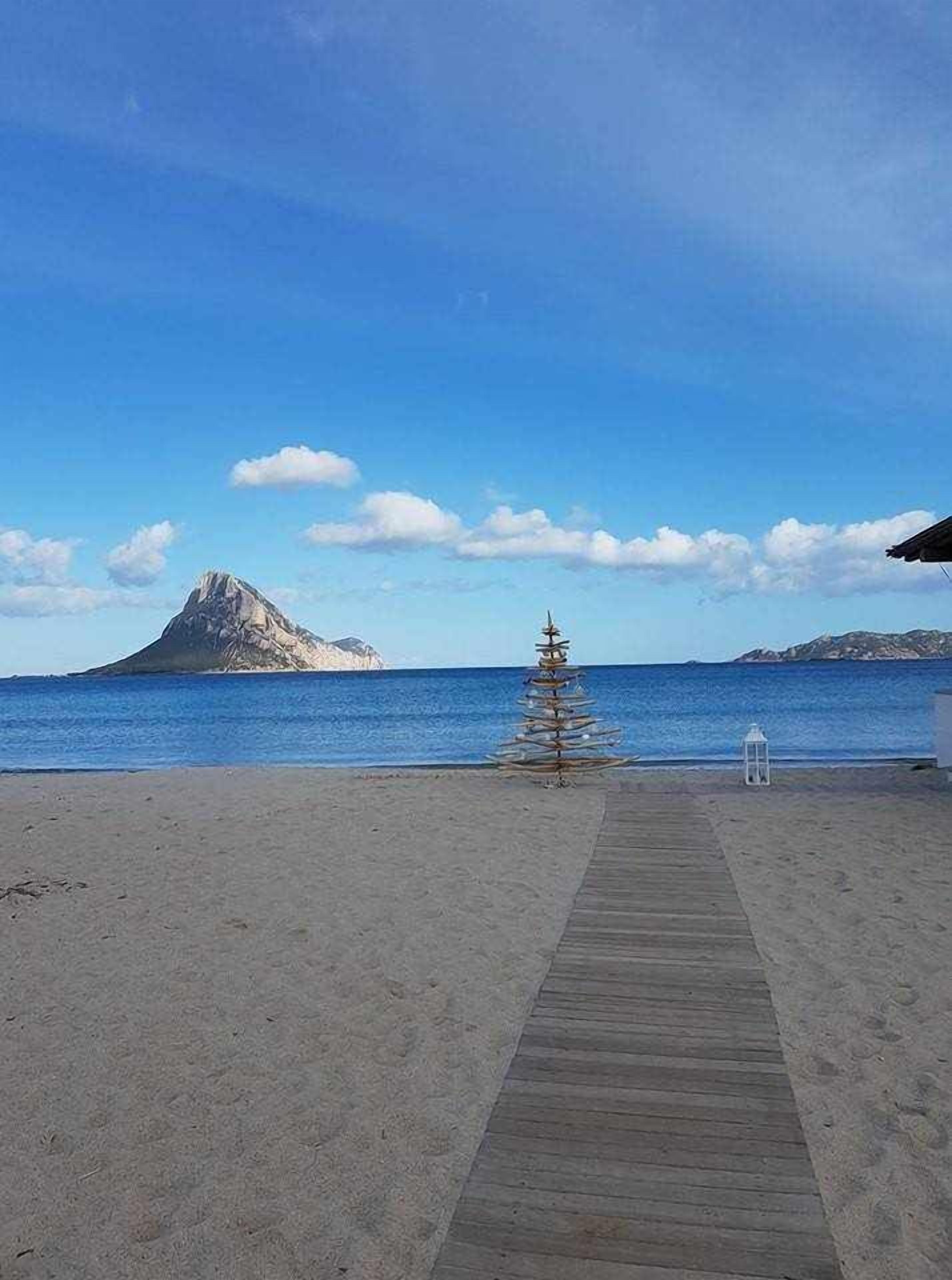 A wooden pathway leading to a beach with a small Christmas tree and lantern, overlooking a calm sea and a mountain.