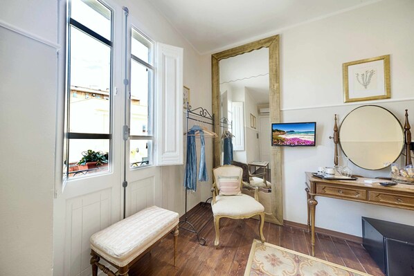 Une pièce comprenant un lit, une chaise, une commode, un miroir et une fenêtre donnant sur des immeubles.