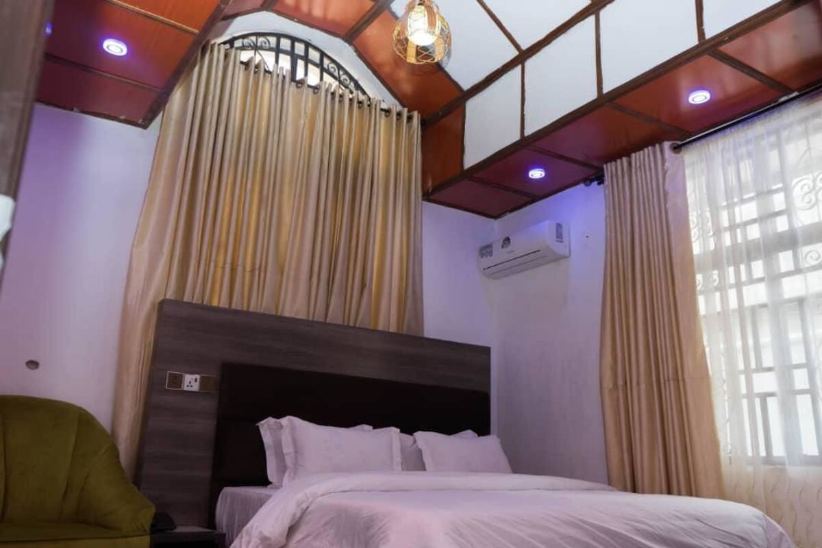 Una habitació d'hotel amb un llit, un butac armchair verd, una làmpada al sostre i una finestra amb cortines.