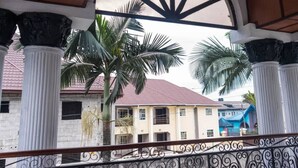 A covered balcony with ornate railings, palm trees, and a building with a red roof in the background. - SS3 Boutique Hotel (Port Harcourt)