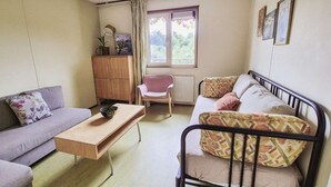 A small, cozy room with a sofa, a chair, a bed, a wooden table, and a window with curtains.
