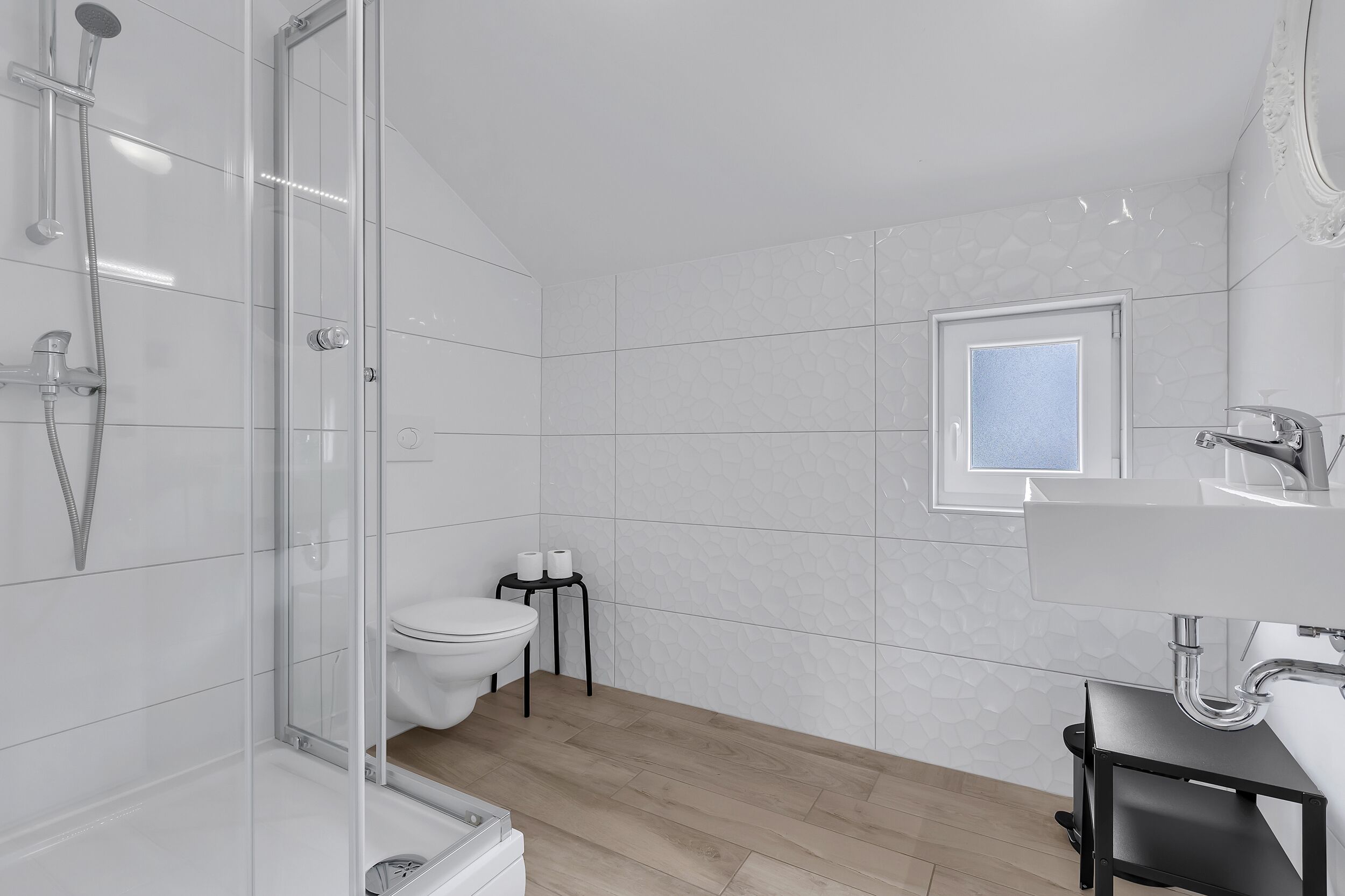 Un baño moderno con ducha envuelta en vidrio, inodoro, ventana pequeña y lavamanos con encimera negra.