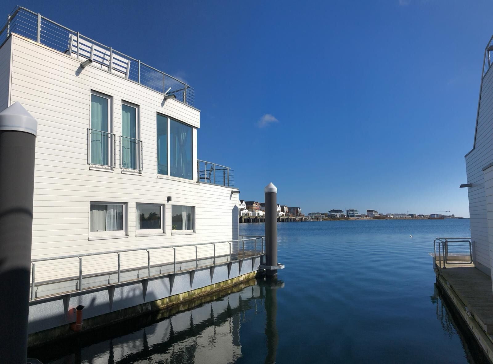 Een wit gebouw met grote ramen en een balkon, afgemeerd aan een steiger met kalm water en een heldere lucht.