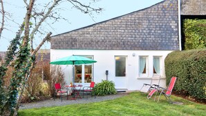 A modern house with a white exterior, a tiled roof, and a small patio area. There is a green umbrella, a red chair, and a white chair on the patio. - Cottage of Suzette ---- (LA FORET-FOUESNANT)