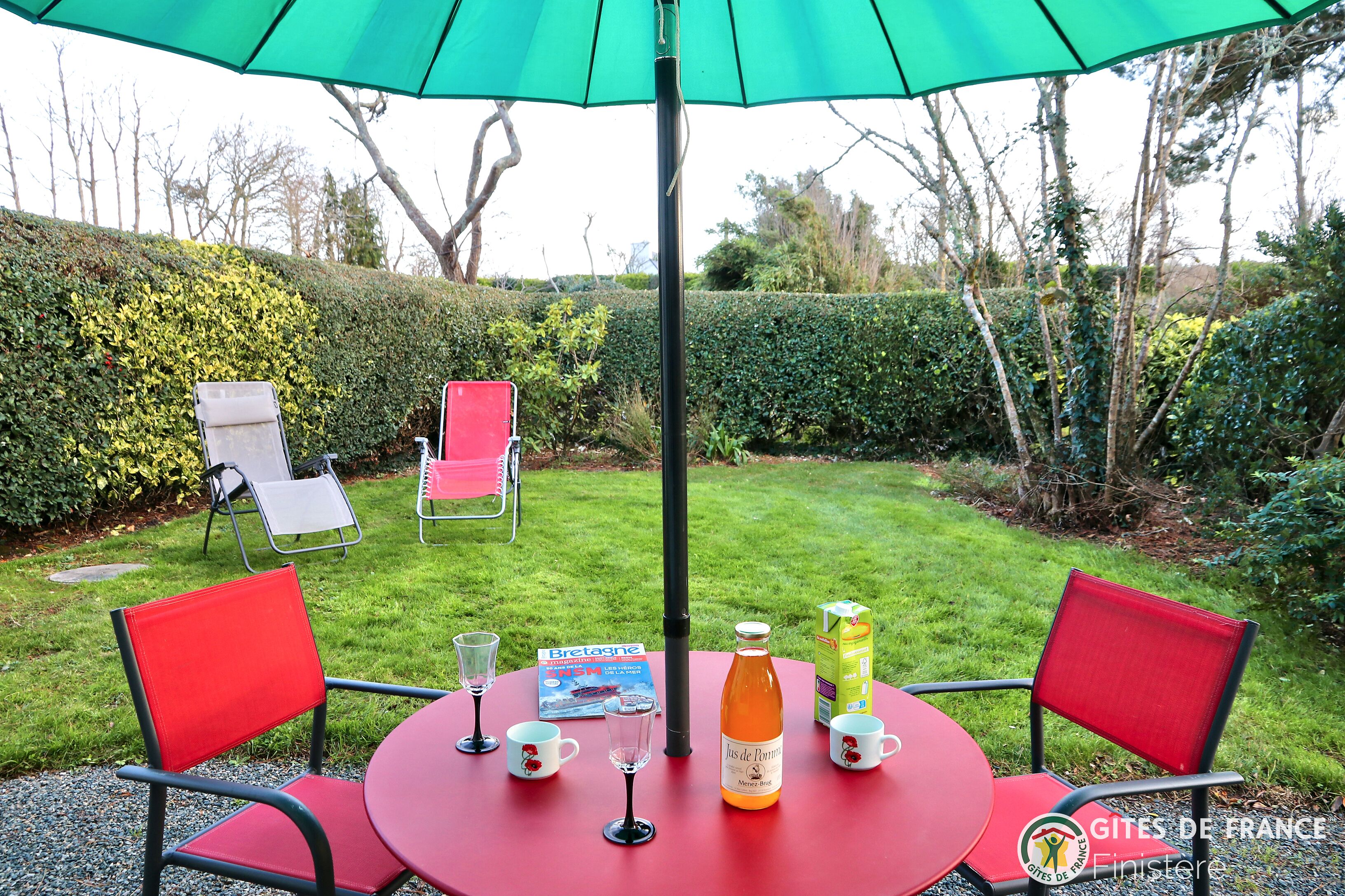 An outdoor seating area with a round table, red chairs, a green umbrella, and a bottle of wine.