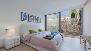 A modern bedroom with a large bed, a bedside table, a lamp, and a view of a stone wall and greenery outside.