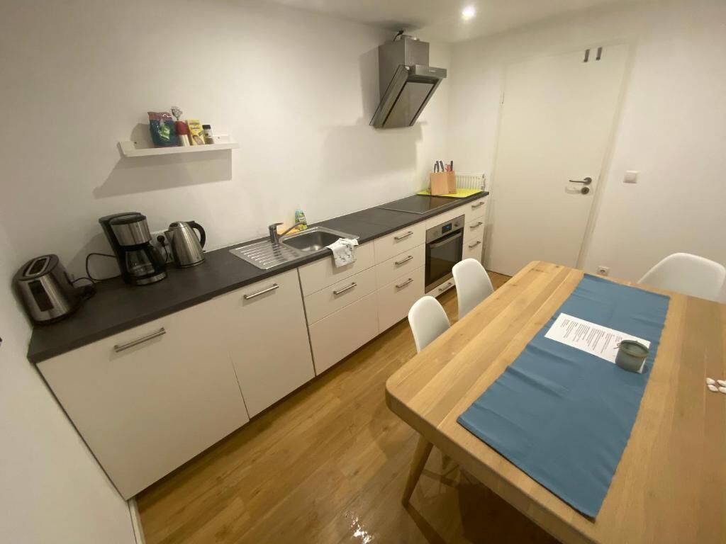 Une cuisine compacte équipée d’une machine à café, d’une bouilloire et d’un évier. On y trouve une table à manger en bois, avec un set de table et une chaise.