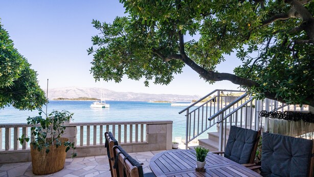 Eine Terrasse mit Tisch und Stühlen, mit Blick auf ein Gewässer mit einem Segelboot.