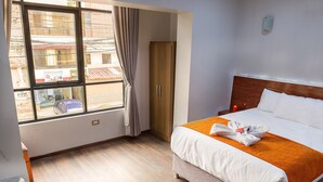 Ein Hotelzimmer mit Bett, Holzboden, einem Fenster mit Vorhängen, einem Kleiderschrank und einem Nachttisch mit Kerze.