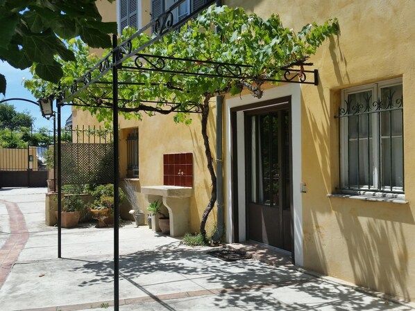 Un edificio con fachada amarilla, una pérgola metálica cubierta de hiedra, una fuente de agua con azulejos y una puerta con ventanal.