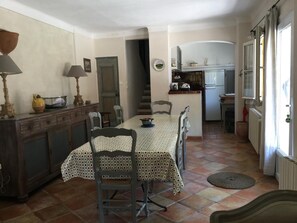 Un comedor con una mesa dispuesta para cuatro personas, rodeada de sillas. Se ve una cocina al fondo con un refrigerador y una estufa.
