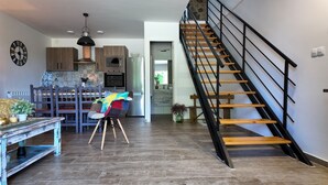 Eine moderne Küche mit Holzausbau, einem Einbauofen und einem Essbereich mit Holztisch und -stühlen. Eine Treppe mit hölzernen Stufen und einem schwarzen Geländer führt auf eine erhöhte Ebene.