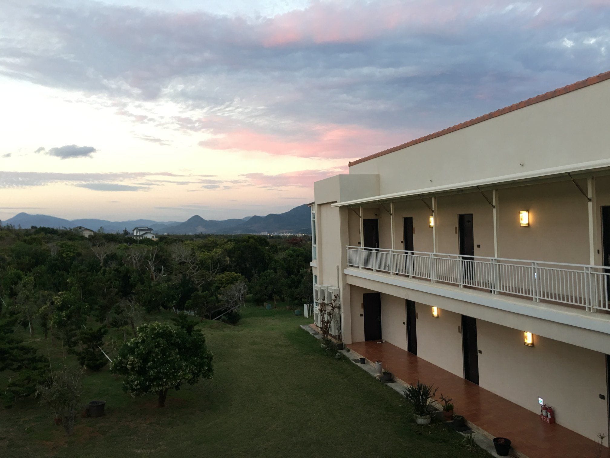 A two-story building with a balcony on the upper level, surrounded by greenery and mountains in the background.