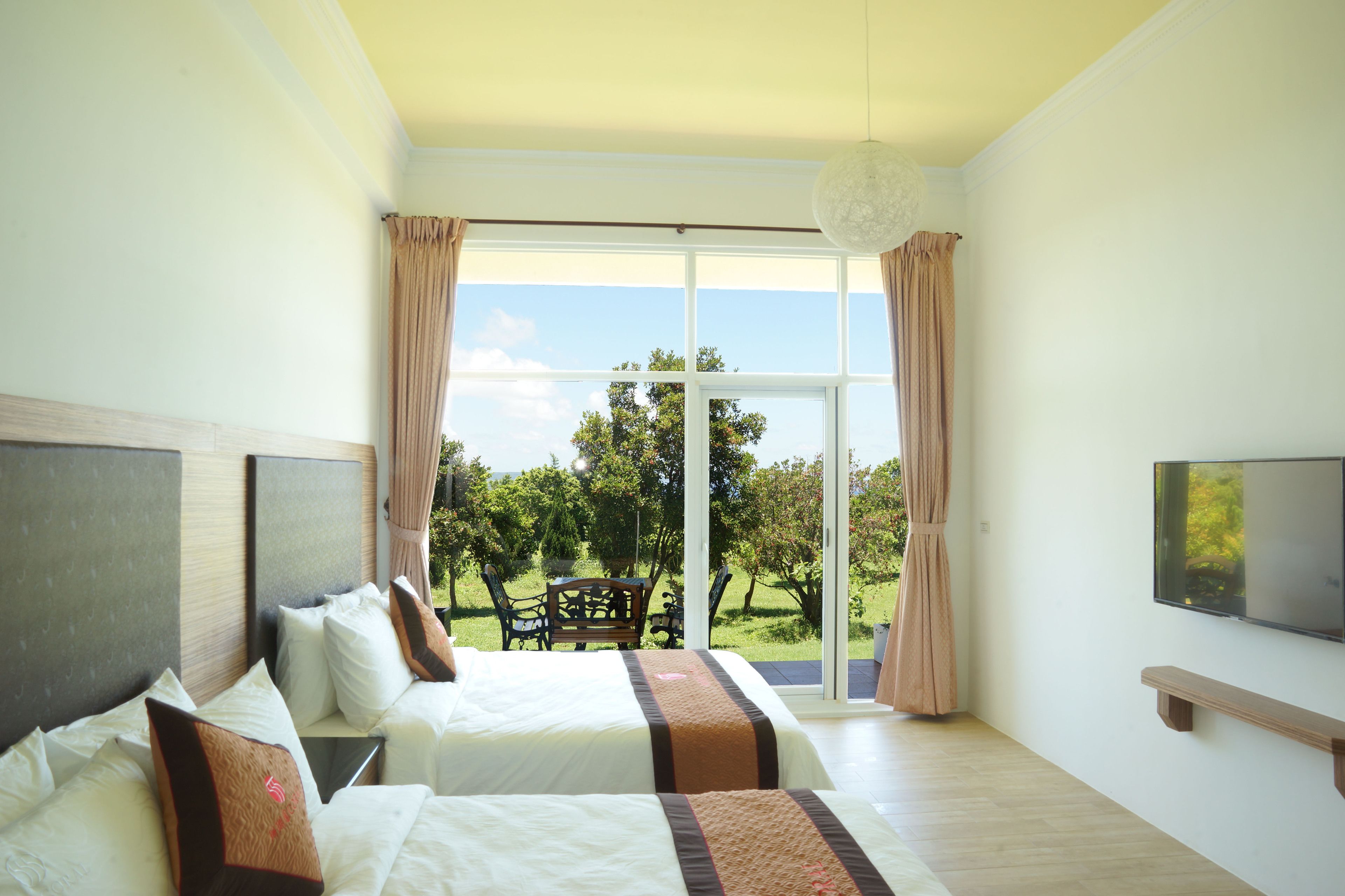 A hotel room with two beds, a TV, and a balcony with a view of trees.