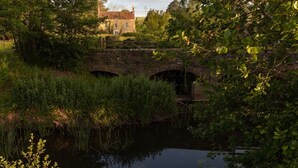 Un pont de pierre enjambant une rivière paisible, entouré d’une végétation luxuriante et avec un bâtiment visible au loin.