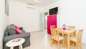 A compact living space with a grey sofa, a small round table, a wooden dining table with chairs, a pink cabinet, and a flat-screen TV mounted on the wall.