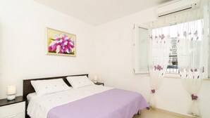 A bedroom with a bed, nightstands, a window with sheer curtains, and a wall-mounted air conditioning unit.