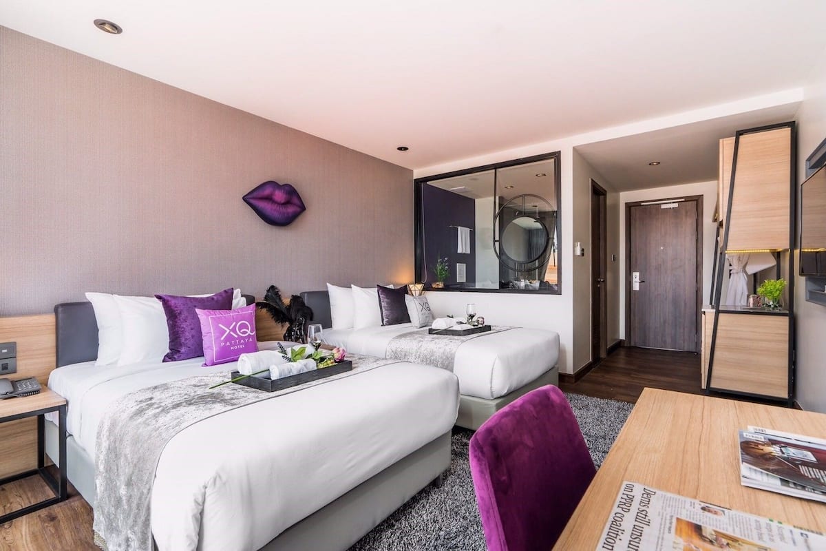 A modern hotel room with two beds, a large mirror, a wooden desk, and a purple chair.